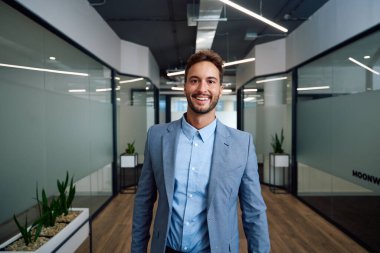 İş kıyafetleri giyen kendine güvenen beyaz bir adam şirket ofisinin koridorunda kameraya bakıyor.