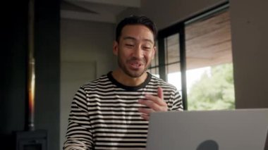 Penceresi perdeli bir odada sakallı bir adam dizüstü bilgisayar kullanırken gülümsüyor ve eğleniyor. Gözlük takıyor ve yakınlarda ses ekipmanı ve cam var.