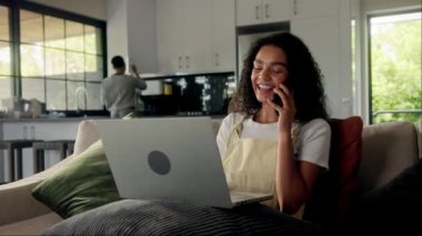 Bir kadın rahat bir şekilde iyi aydınlatılmış bir odada koltukta oturuyor, dizüstü bilgisayar kullanıyor ve cep telefonuyla konuşuyor. Binaların pencereleri doğal ışığa izin veriyor, rahat bir atmosfer yaratıyor.
