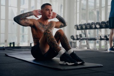 Üstsüz, genç, dövmeli, şort giyen spor salonunun paspasında mekik çeken adam.