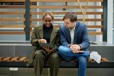 İş kıyafetleri giyen iki yetişkin koridorda bankta dijital tablet kullanırken oturup konuşuyorlar.