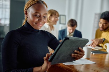 Ofisteki konferans masasında dijital tablet kullanarak iş kıyafetleri giyen dört genç çok ırklı yetişkin.