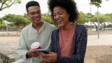 Karışık ırk çifti parkta takılırken bankta telefona gülüyor. Yüksek kalite 4k görüntü