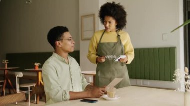 Afrika kökenli Amerikalı garson modern bir kahve dükkanındaki not defterine karışık bir ırk müşterisi siparişi veriyor. Yüksek kalite 4k görüntü