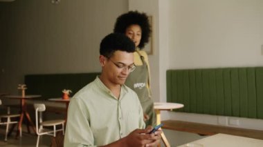 Karışık bir yarış müşterisi, modern bir kafede garsonun siparişini almasını beklerken telefonuyla mesajlaşıyor. Yüksek kalite 4k görüntü