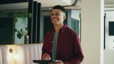 Gülümseyen genç işadamı modern ofiste yürürken tablet kullanıyor.