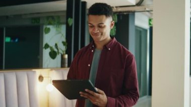 Gündüz vakti modern ofiste yürürken tablet bilgisayar kullanarak akıllı ve rahat bir şekilde gülümseyen genç işadamı.