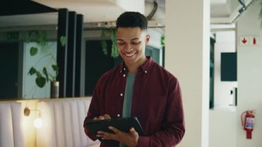 Gündüz vakti modern ofiste yürürken tablet bilgisayar kullanarak akıllı ve rahat bir şekilde gülümseyen genç işadamı.