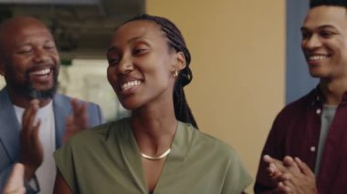 Bir grup farklı iş arkadaşı, gündüz vakti parlak bir ofis alanında siyahi iş kadınını alkışlıyor.