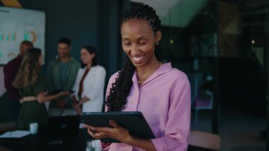 Modern ofiste tablet kullanan gülümseyen genç iş kadını ve arka planda meslektaşları.