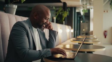 Modern ofis salonunda dizüstü bilgisayar kullanarak gülümseyen olgun, siyah iş adamının yan görüntüsü.