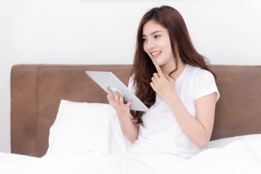 Güzel Asyalı kadın beyaz yatak odasındaki yatakta tableti kullanıyor.