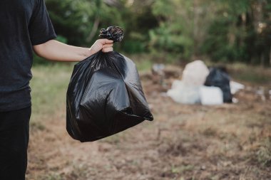 Elinde plastik çöp torbası olan Asyalı bir adam, arka planda çöp yığını, çevre sorunları ve sürdürülebilirlik kavramı.