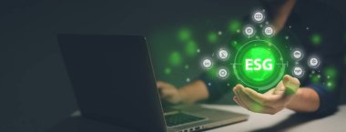 Dizüstü bilgisayar kullanan bir adam bir dünyanın hologramına dokunuyor. Dünya genelinde ESG kavramını temsil eden ikonlar var, bu kavram çevre, sosyal ve yönetim anlamına geliyor. Adam ESG kavramını keşfediyor ve bunun önemini daha iyi anlıyor.