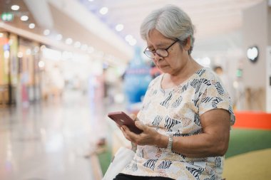 Alışveriş merkezinde otururken akıllı telefonunu kullanan mutlu bir Asyalı kadın. Gülümsüyor ve telefonuyla meşgul görünüyor. Arkaplan bulanık, ve resim orta ekran ile çekiliyor.