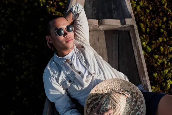 An Asian man finds peace in the heart of nature, relaxing on a wooden boat surrounded by lush greenery. This image captures the essence of an active lifestyle, relaxation, travel, and connection with nature, highlighting the beauty of local culture i