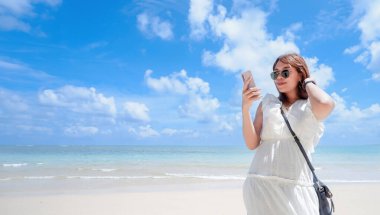 Tropik bir plajda, akıllı telefonuna takılmış bir kadın. Açık mavi su ve gökyüzü sakin bir zemin oluşturuyor, günlük hayatımızdaki teknoloji ve rahatlığın karışımını vurguluyor..