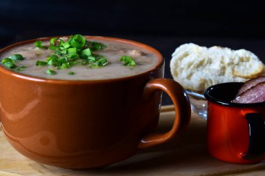 Frenk soğanlı fasulye çorbası, pastırma ve ekmek, ahşap bir tabanda bir toprak kabında servis edilir.