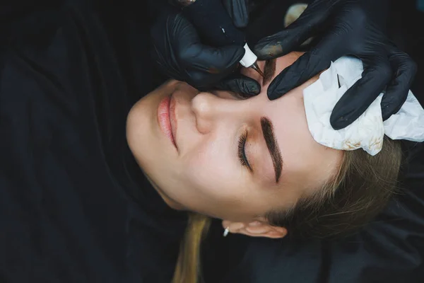 Beautician hand doing brow permanent makeup on an attractive female face