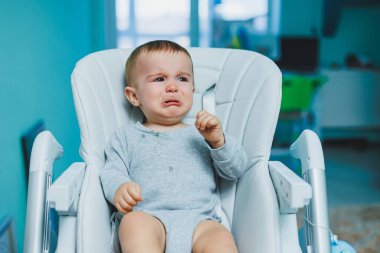 Küçük bir çocuk yüksek bir sandalyede oturuyor ve ağlıyor. Beslenmek için bebek koltuğu. Küçük bir çocuk oturuyor. Çocukların duyguları.