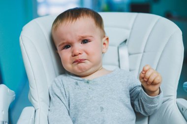 Küçük bir çocuk yüksek bir sandalyede oturuyor ve ağlıyor. Beslenmek için bebek koltuğu. Küçük bir çocuk oturuyor. Çocukların duyguları.