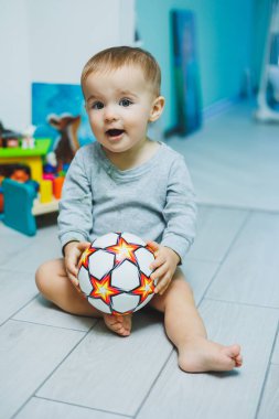 Sarışın küçük bir çocuk, çıplak ayaklarıyla bir odada yerde oturuyor. Surat asıyor ve elinde turuncu bir balon tutuyor.