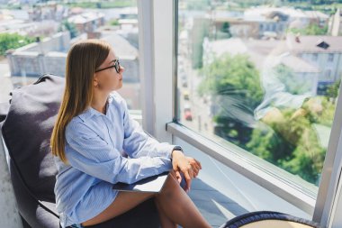 Gözlüklü genç bir kadın pencerenin yanındaki yumuşak bir divanda oturur ve gökdelenden şehre bakar. Uzak çalışma