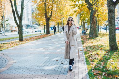 Sonbahar parkında yürüyen, akıllı telefondan konuşan, trençkot giyen, güneş gözlüğü takan, mutlu ruh hali, moda tarzı eğilimi olan genç bir kadın.
