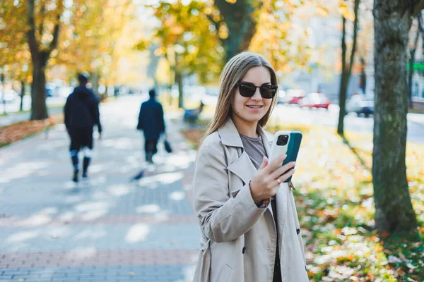 stock image A beautiful woman in sunglasses is walking in the autumn park and talking on the phone. Joyful girl on the autumn alley in the city.