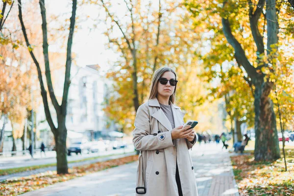 秋の公園を歩く若い笑顔の女性 スマートフォンで話して トレンチコートを着て サングラス 幸せな気分 ファッションスタイルの傾向 — ストック写真