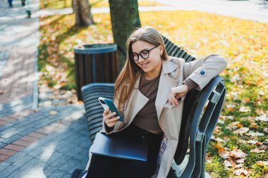 Gülümseyen bir kadın Autumn Park 'taki bankta dizüstü bilgisayarla çalışıyor. Harika bir gülüşü, uzun saçları ve büyük mavi gözleri var. Modern çalışan bir kadının portresi. Sarı park arkaplanı