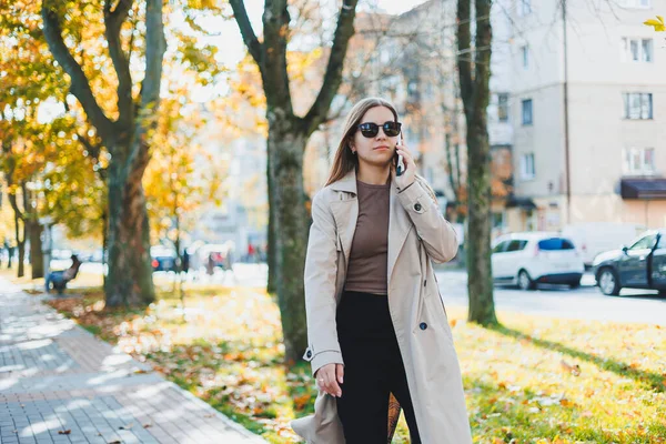 Ung Leende Kvinna Promenader Höstparken Talar Smartphone Bär Trenchcoat Solglasögon — Stockfoto