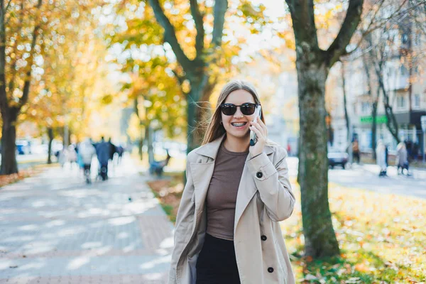 秋のコートを着たスタイリッシュな幸せな女性とサングラスが屋外を歩き 携帯電話で話し 陽気な女性が公園を歩いている間に屋外の携帯電話で話してチャットします — ストック写真