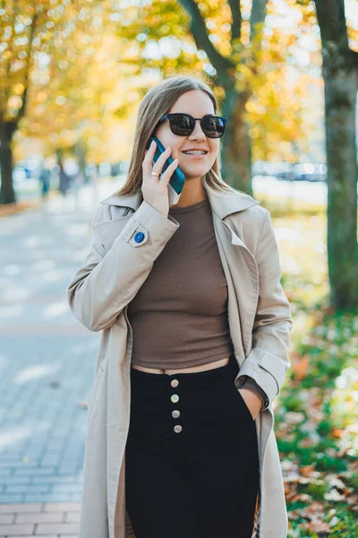 秋のコートを着たスタイリッシュな幸せな女性とサングラスが屋外を歩き 携帯電話で話し 陽気な女性が公園を歩いている間に屋外の携帯電話で話してチャットします — ストック写真