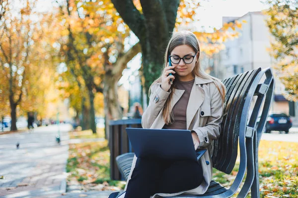 stock image A smiling woman works with a laptop on a bench in the Autumn Park. She has a great smile, long hair and big blue eyes. Portrait of a modern working woman. Yellow park background