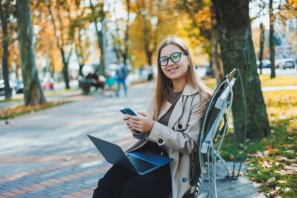 笑顔の女性は 秋の公園のベンチでラップトップで動作します 彼女は大きな笑顔 長い髪と大きな青い目をしています 現代の働く女性の肖像画 黄公園の背景 — ストック写真