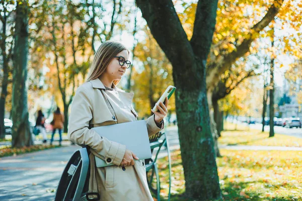 ノートパソコンや携帯電話を使って遠隔作業をする秋のコートの美しい若い女性 秋の公園で携帯電話で話す女性 — ストック写真