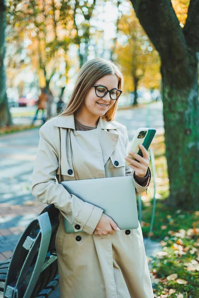 Beautiful young woman in autumn coat using laptop and phone working remotely, woman talking on mobile phone in autumn park.