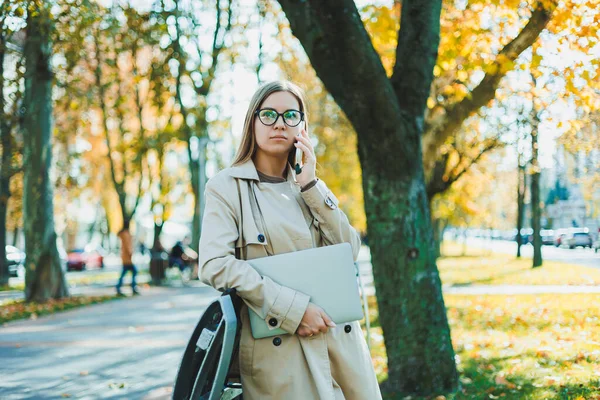 ノートパソコンや携帯電話を使って遠隔作業をする秋のコートの美しい若い女性 秋の公園で携帯電話で話す女性 — ストック写真