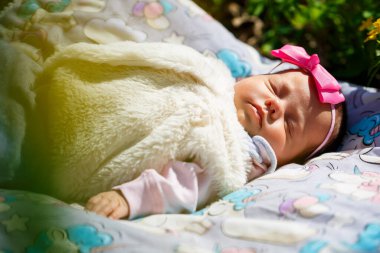 Başında fiyonk olan yakın plan bir bebek yeşil çimlerin üzerinde beyaz bir ekosenin üzerinde yatıyor. Bahar sokakta, güneş çocuğun üzerinde parlıyor.