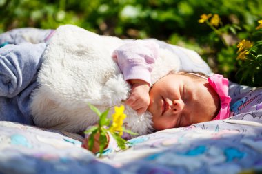 Başında fiyonk olan yakın plan bir bebek yeşil çimlerin üzerinde beyaz bir ekosenin üzerinde yatıyor. Bahar sokakta, güneş çocuğun üzerinde parlıyor.