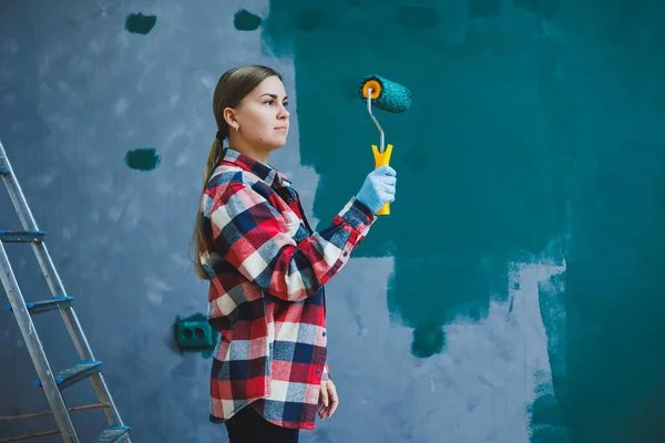 stock image A young beautiful woman designer makes repairs, paints a gray wall with green paint with a roller. Plaid shirt, long hair and jeans.