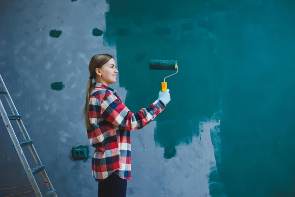 stock image A young beautiful woman designer makes repairs, paints a gray wall with green paint with a roller. Plaid shirt, long hair and jeans.