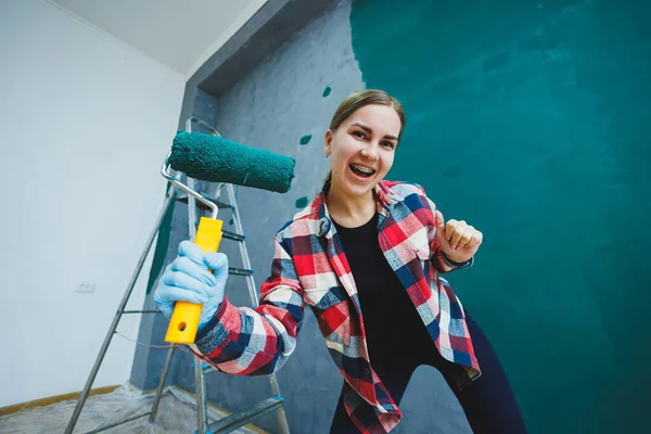 An emotional woman paints the walls. Young smiling pretty woman in shirt holding roller in right hand and singing. Repair in the room.