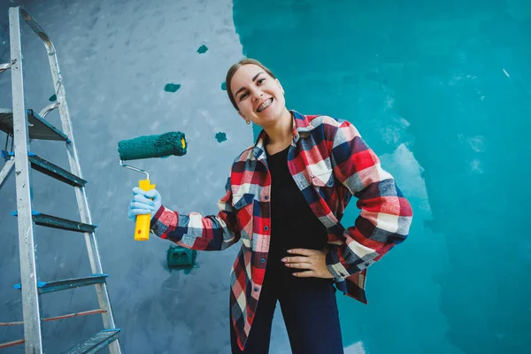 An emotional woman paints the walls. Young smiling pretty woman in shirt holding roller in right hand and singing. Repair in the room.