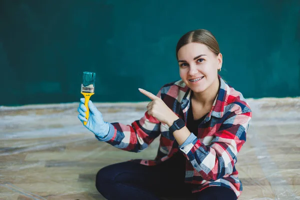 A beautiful woman designer makes repairs, paints a gray wall with green paint with a brush. Plaid shirt, long hair and jeans. Repair in the apartment