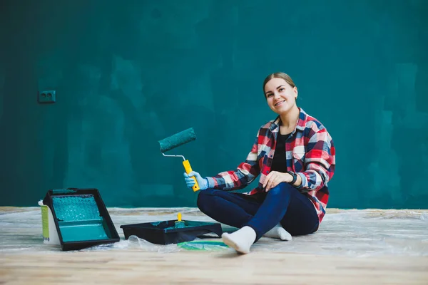 A beautiful woman designer makes repairs, paints a gray wall with green paint with a brush. Plaid shirt, long hair and jeans. Repair in the apartment