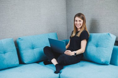 Genç bir kadın kanepede oturuyor ve bir dizüstü bilgisayar üzerinde çalışıyor. Uzaktan kumanda. Evde internet üzerinden çalış.