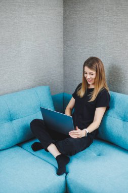 Genç bir kadın kanepede oturuyor ve bir dizüstü bilgisayar üzerinde çalışıyor. Uzaktan kumanda. Evde internet üzerinden çalış.