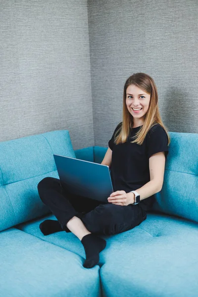 stock image A young woman is sitting on a sofa and working on a laptop. Remote work. Work at home online